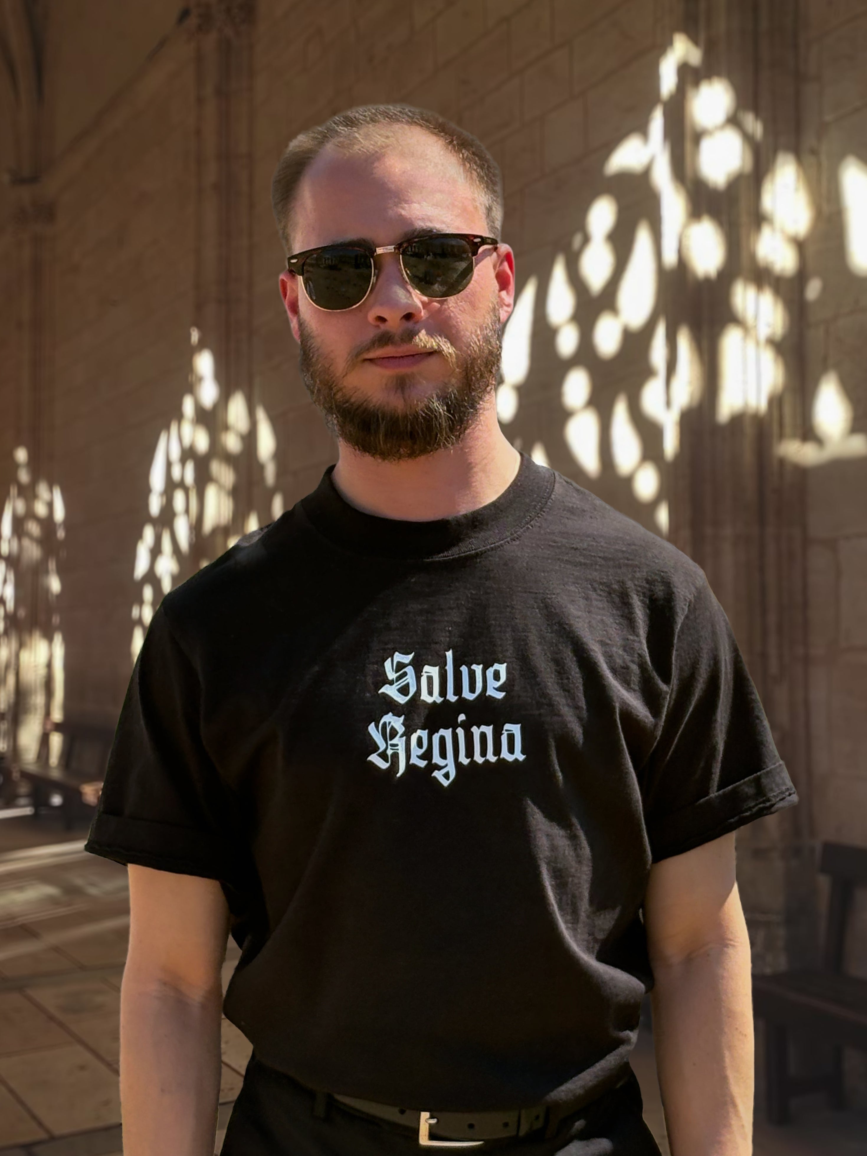 Man wearing a black t-shirt with 'Salve Regina' text, standing in a sunlit architectural setting.
